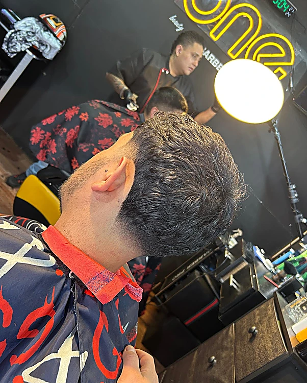 Corte terminado visto de espaldas; al fondo, un barbero de One trabaja bajo el neón ámbar del letrero
