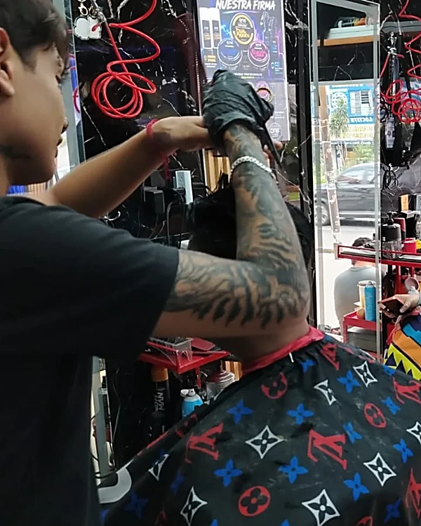 Barbero con el brazo tatuado y guantes negros cortando con tijera a un cliente de espaldas con capa estampada