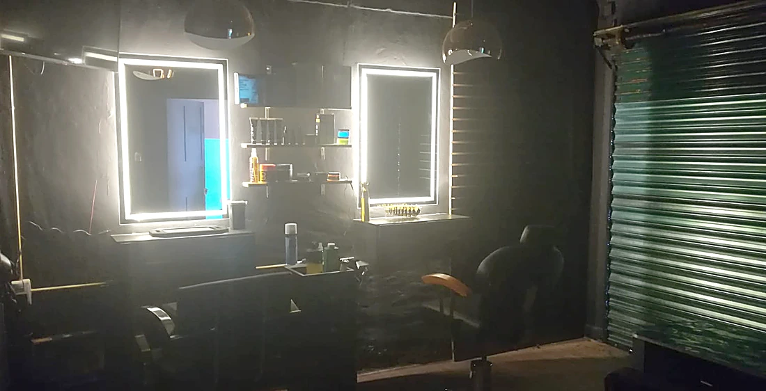 Dos estaciones de corte con espejos de contorno de luz blanca sobre pared negra, sillones y la persiana verde de One Barber Boutique