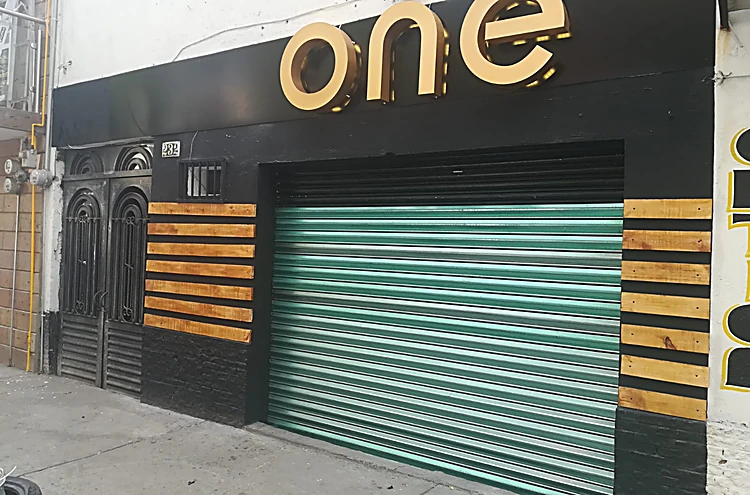 Fachada de día: letras one doradas sobre muro negro, listones de madera y persiana verde-azulada