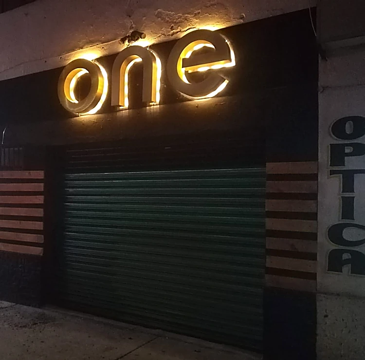 Fachada de noche con las letras one iluminadas en ámbar sobre fondo negro, listones de madera y persiana verde