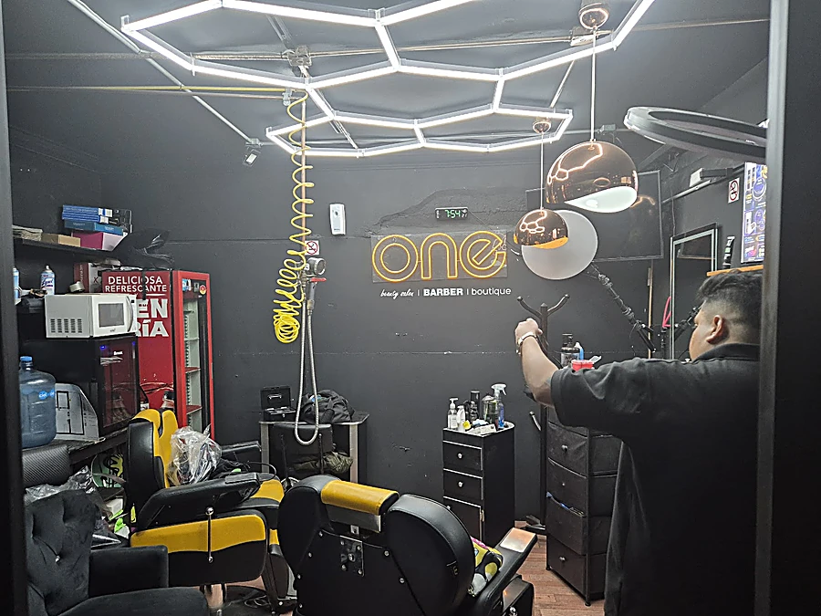 Interior de One: techo de hexágonos de luz LED blanca, letrero de neón ámbar one beauty salon barber boutique, sillones negros con amarillo y un barbero al fondo