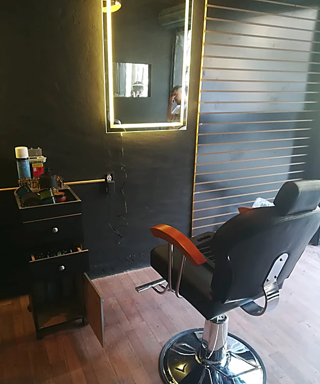 Silla de barbero negra con brazos de madera frente a un espejo con contorno de luz, en un muro negro